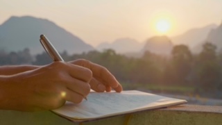 Young woman writing travel notes in diary during sunrise with beautiful sun light and mountain landscape on the background, close-up.